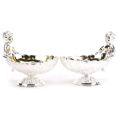 1208 - Pair of silver plated open table salts with spoons in the form of Putti holding a shell, 12cm in len... 