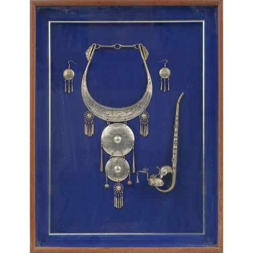 1310 - Thai white metal jewellery and opium pipe housed in a glazed framed display, overall 55cm x 42.5cm