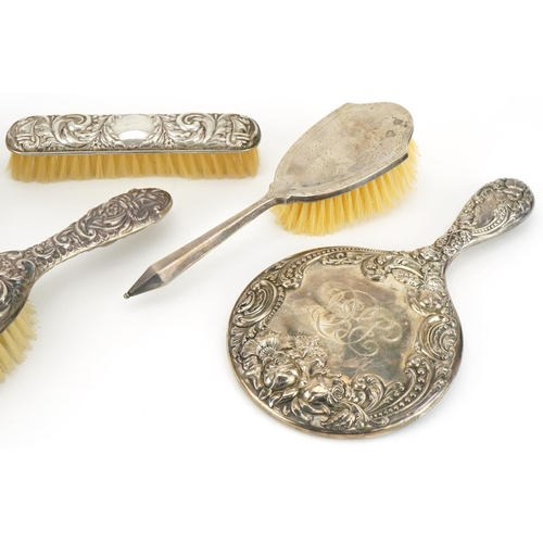 2446 - Four silver backed vanity brushes and a hand mirror, four profusely embossed with foliage, the large... 