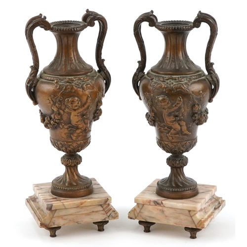 36 - Pair of patinated spelter twin handled urns embossed with Putti, raised marble bases, 36cm high.