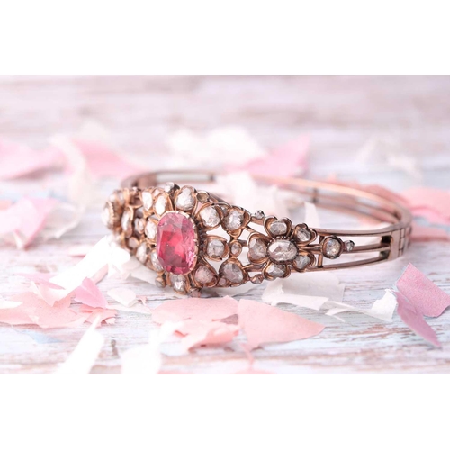 72 - An antique yellow metal and diamond bangle, set with a reddish pink central stone, likely spinel, th... 