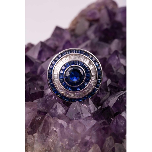 127 - A sapphire and diamond cluster ring; the central circular cut sapphire in rubover mount within conic... 