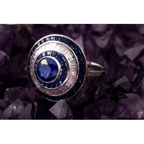 127 - A sapphire and diamond cluster ring; the central circular cut sapphire in rubover mount within conic... 