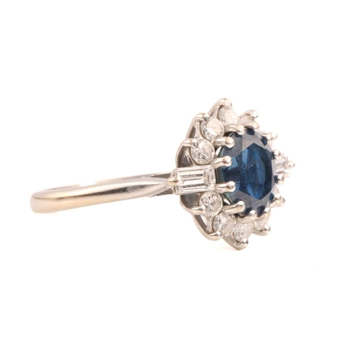 211 - A sapphire and diamond cluster ring in 18ct white gold, consisting of an oval-cut sapphire with deep... 