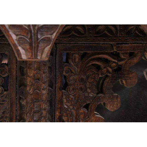 128 - An Indian rustic carved teakwood over mantle mirror with overhanging arcaded cornice above a pair of... 