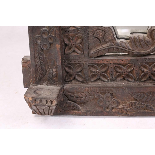 128 - An Indian rustic carved teakwood over mantle mirror with overhanging arcaded cornice above a pair of... 