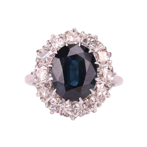 14 - A sapphire and diamond cluster ring, the oval cut sapphire in claw mounts above a border of twelve r... 