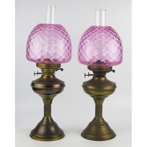 1635 - A pair of brass oil lamps with pink glass shades, circular squat reservoirs raised on reeded columns... 