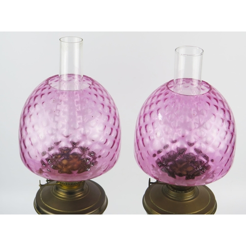 1635 - A pair of brass oil lamps with pink glass shades, circular squat reservoirs raised on reeded columns... 