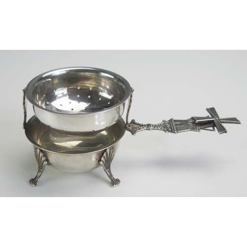 689 - A Dutch white metal tea strainer on stand, the handle with windmill terminal, raised on four swept f... 