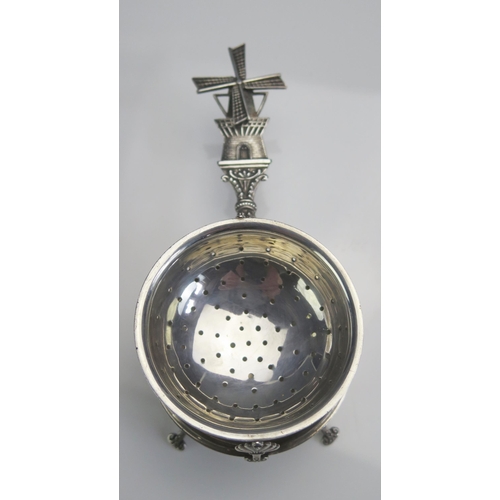 689 - A Dutch white metal tea strainer on stand, the handle with windmill terminal, raised on four swept f... 