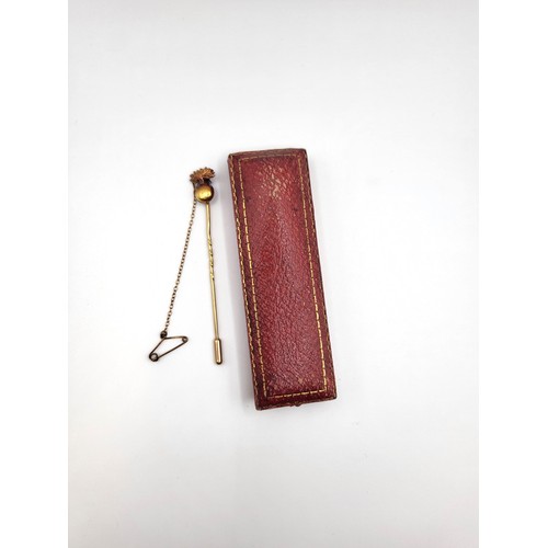 288 - A red leather tooled box with a gold unmarked stick pin with safety chain topped with a motif. 3.3gm... 