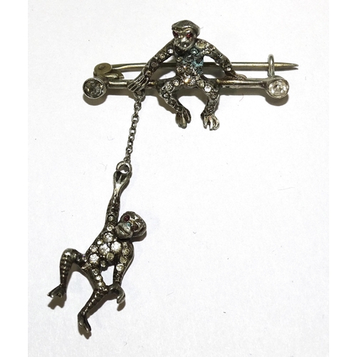 831 - An early-20th century white metal brooch modelled as a monkey sat on a bar with paste terminals, bel... 