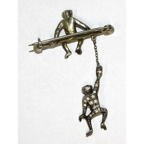 831 - An early-20th century white metal brooch modelled as a monkey sat on a bar with paste terminals, bel... 