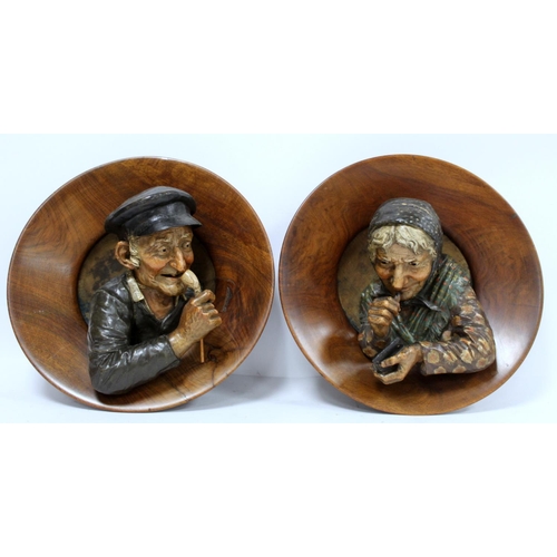 380 - Pair of Continental pottery and treen wall plaques, depicting an elderly man with a pipe and a woman... 