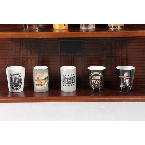 66 - Thirteen John Wayne themed shot glasses, on wall mountable stand.