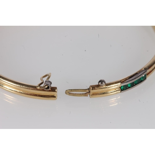 218 - Unmarked yellow gold bangle, on hinged clasp, the face interspersed with channel set round emeralds,... 