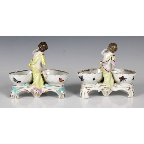 25 - A pair of 20th century KPM, Germany, porcelain salts, each featuring a putti with two baskets in pol... 