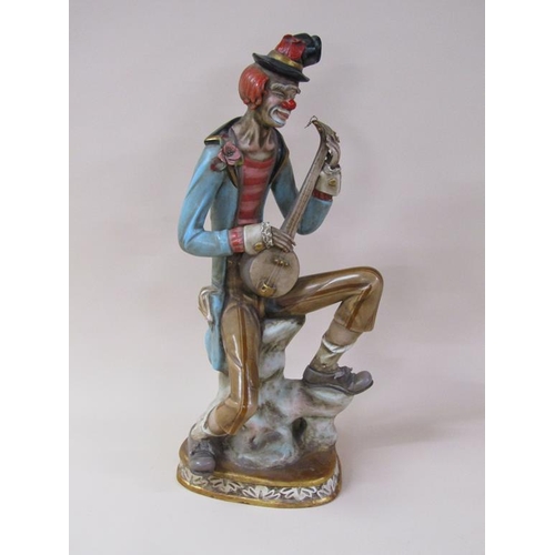 53 - An early 20c painted composite stoneware figure of a clown, seated, playing a string instrument, 81c... 