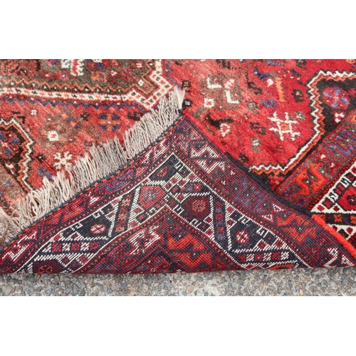 1215 - Persian handwoven wool carpet of red ground, with central diamond design, showing age & wear, cut do... 